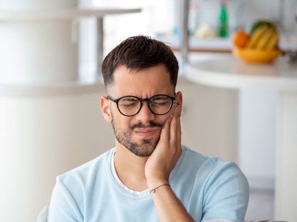Man with glasses sitting in kitchen rubbing jaw in pain