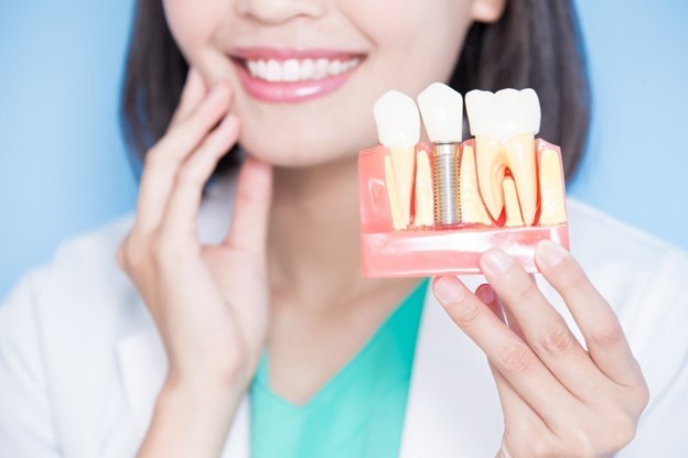 dentist smiling, touching cheek, and holding dental implant model 